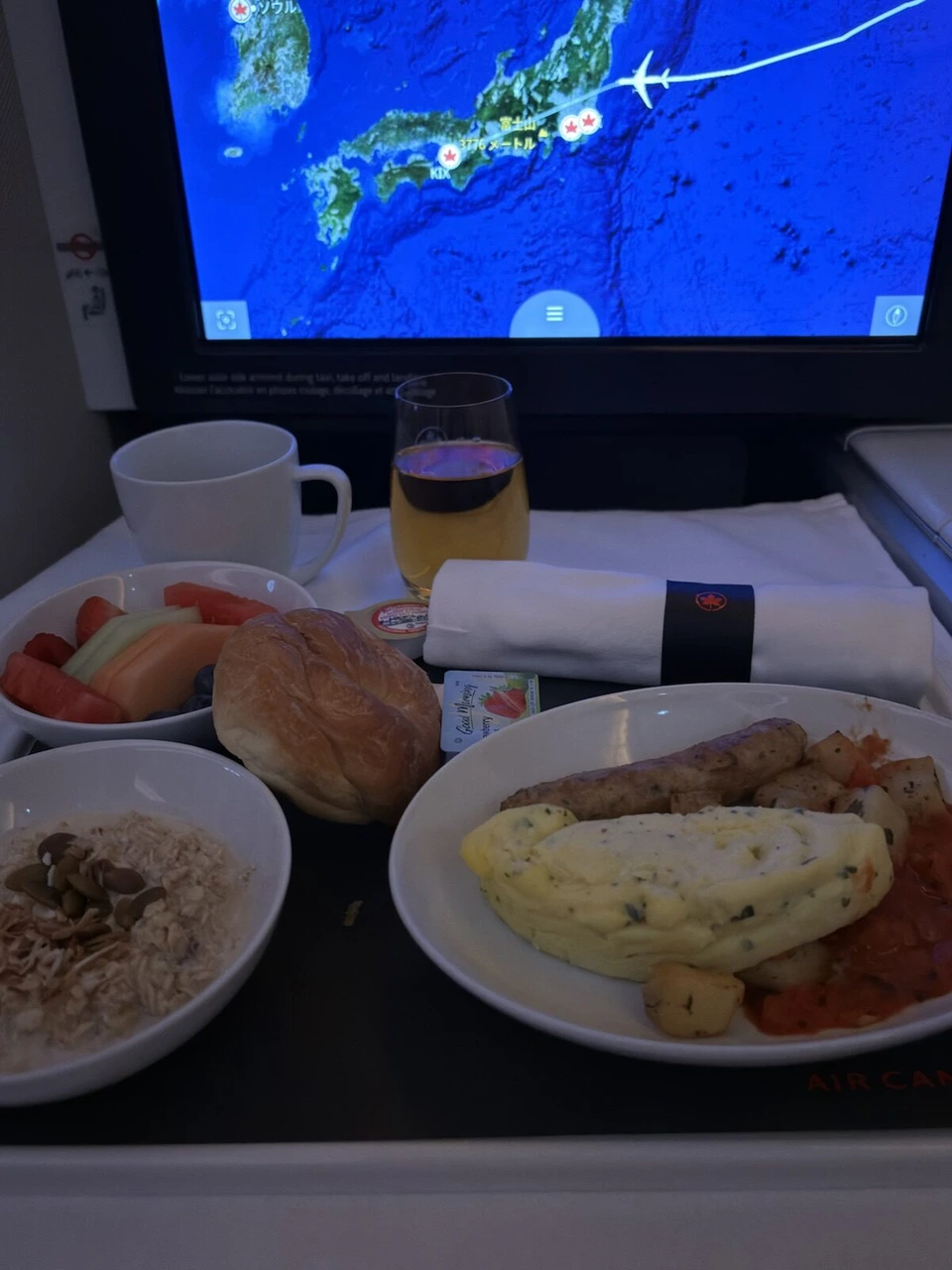 到着前の食事のトレー全体。オムレツとソーセージ、ポテト、オートミール、フルーツ。奥のモニターに日本列島の地図