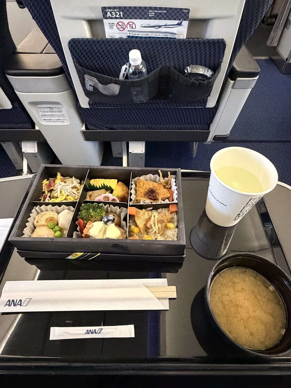 ANA NH578 新千歳→神戸 プレミアムクラスの夕食。仕切りのある黒い弁当箱、味噌汁、紙コップの飲み物。前の座席に「A321」と書かれた安全のしおり