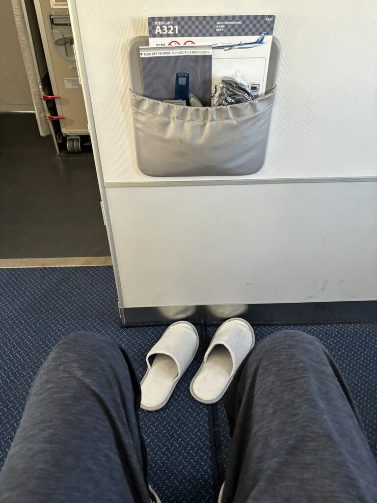 ANA A321 プレミアムクラス最前列1Hの足元。スリッパと、壁のポケットのA321の安全のしおり