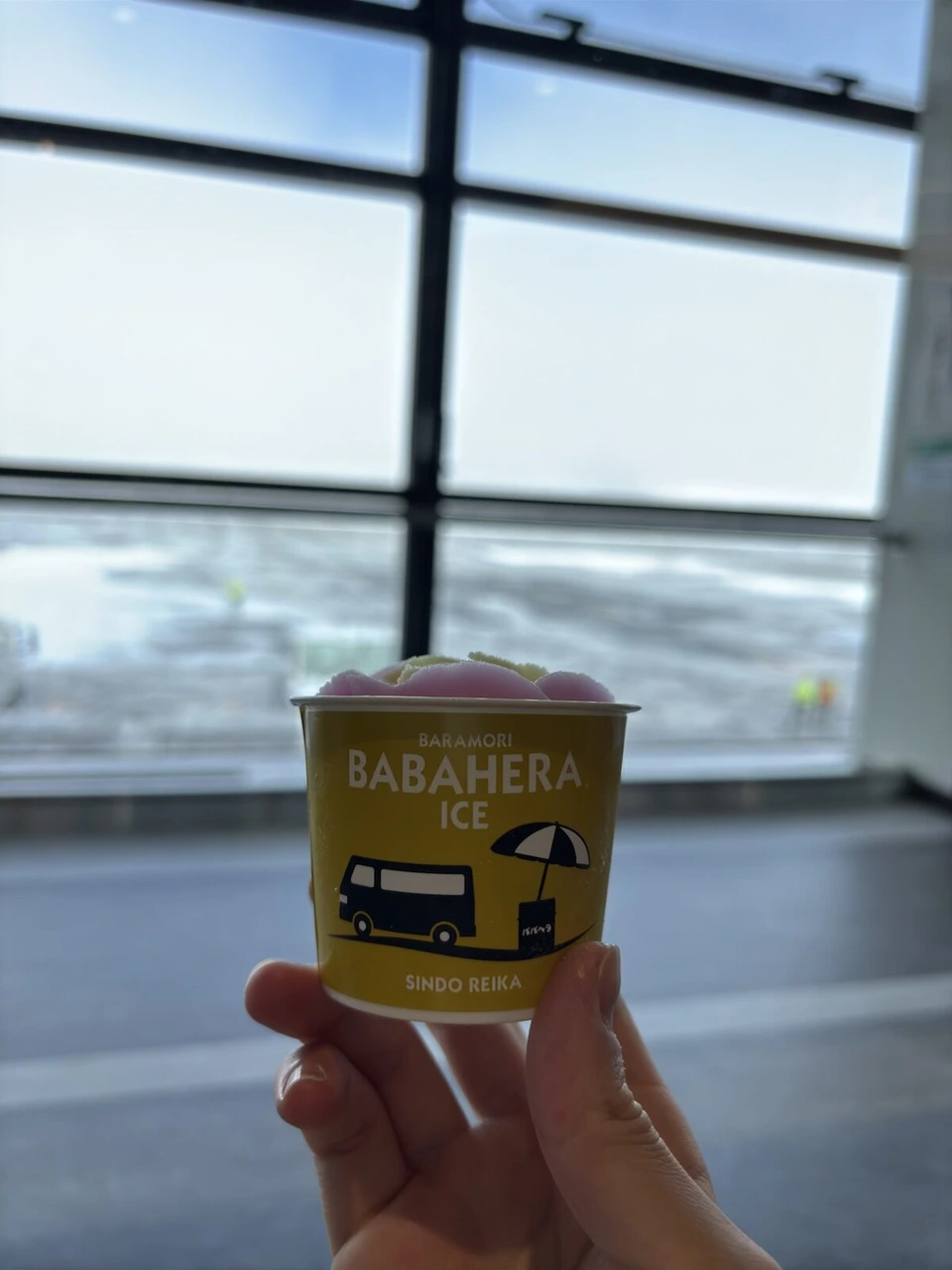 秋田空港の窓際で手に持った、BABAHERA ICEと書かれた黄色いカップのアイス