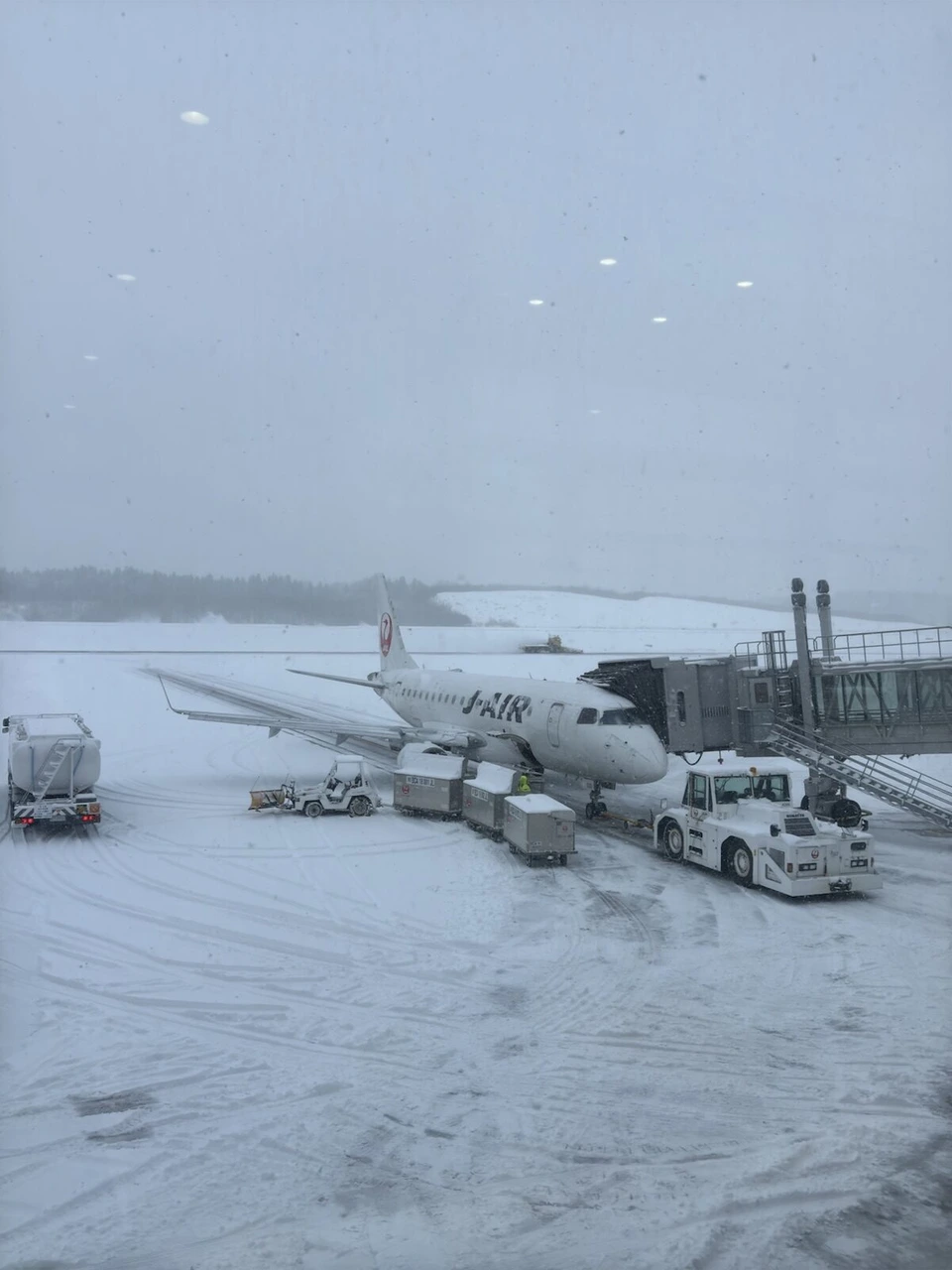 雪が降る秋田空港。ボーディングブリッジにつながるJ-AIRのエンブラエル機と除雪車