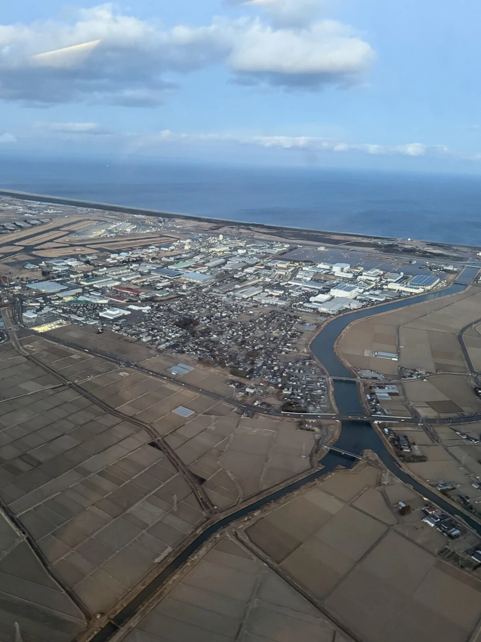仙台空港を離陸した直後の機窓。夕方の光に照らされた川と工場、田んぼ、その先の海