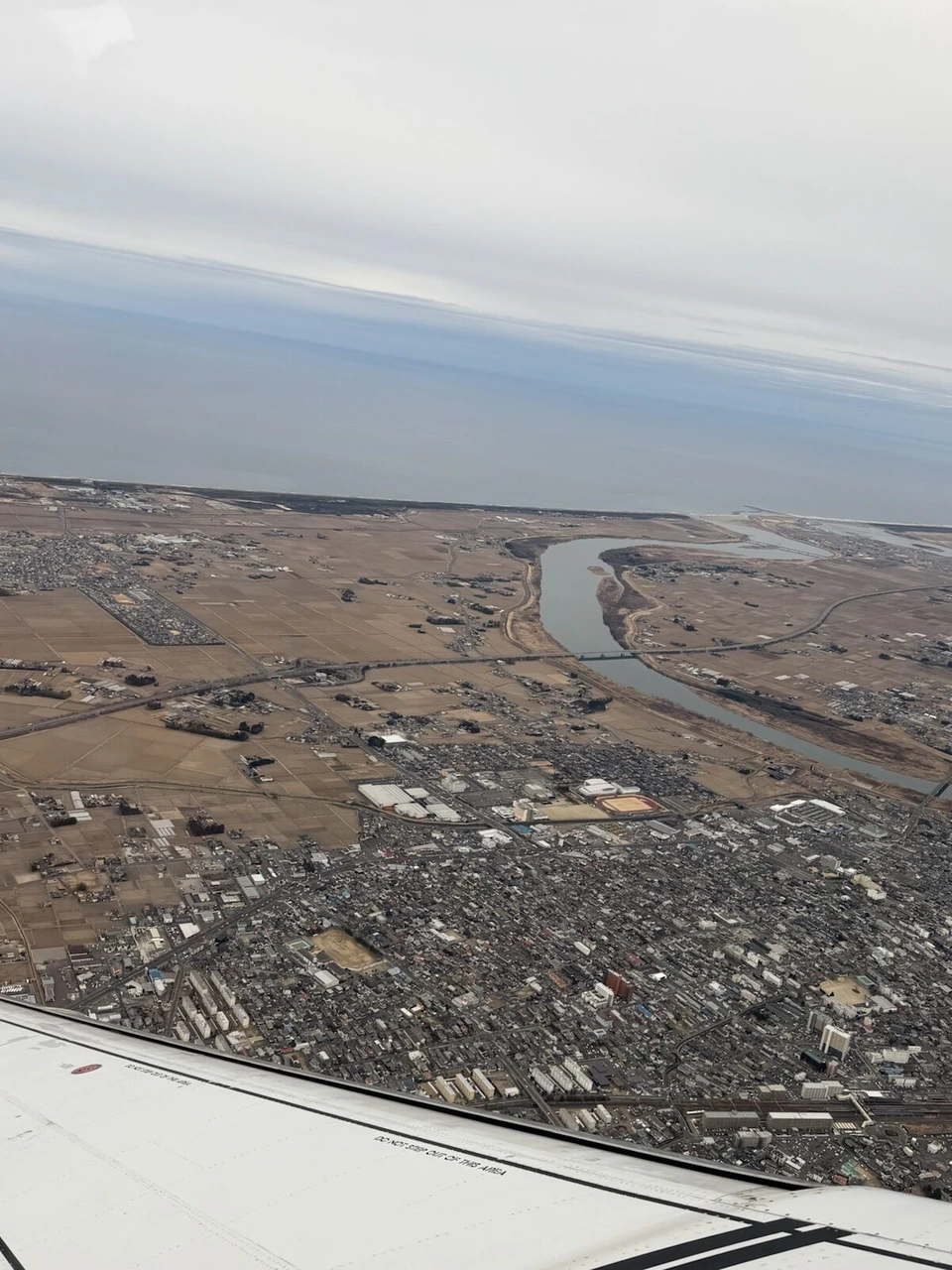 仙台空港を離陸した直後の機窓。曲がりくねった川と田畑、住宅地、遠くに海