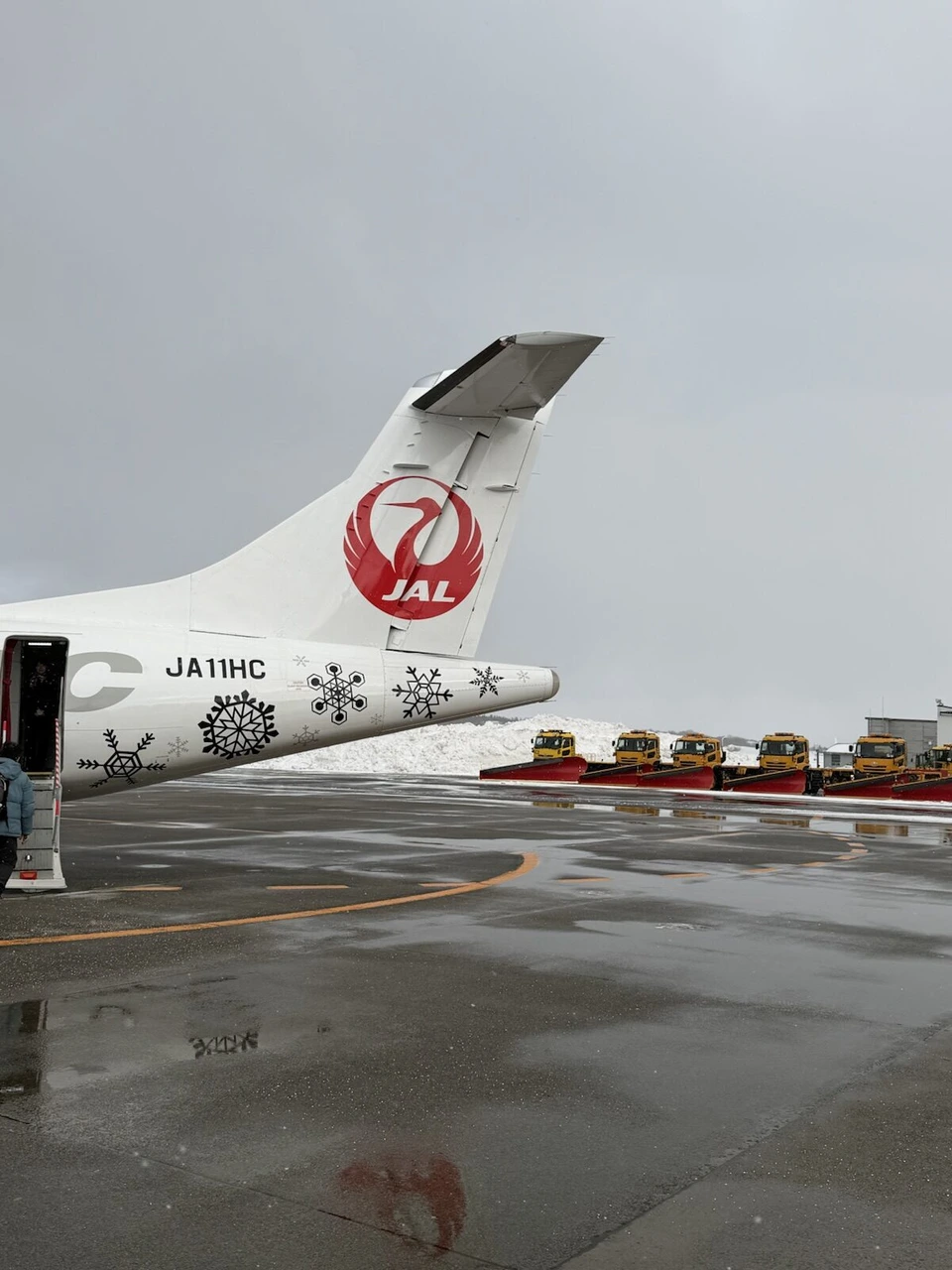 秋田空港の駐機場。雪の結晶が描かれたATR42-600の尾翼とJA11HCの登録記号、奥に並ぶ除雪車