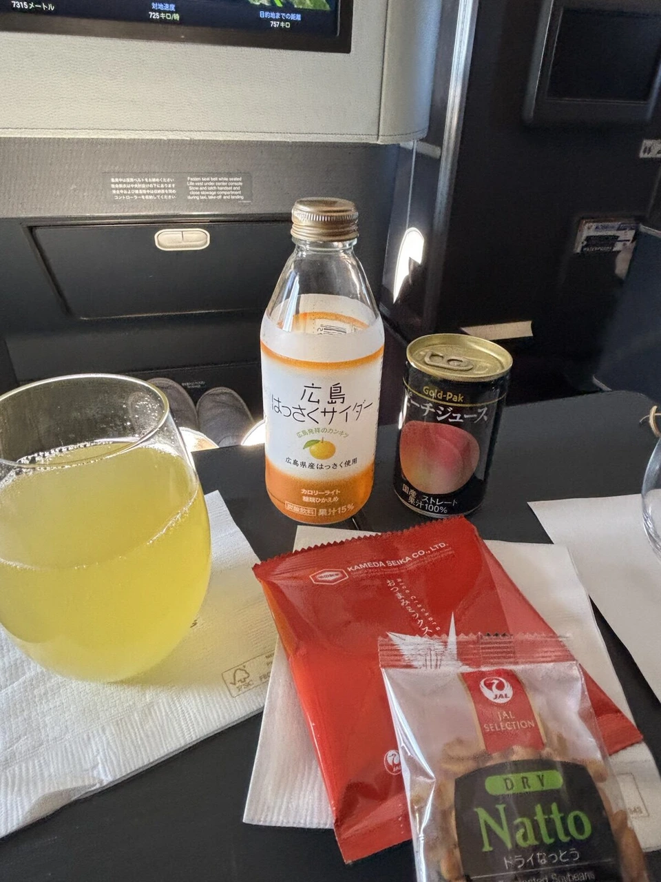 JL2081ファーストクラスのグラスに注いだはっさくサイダーと、赤い袋のお菓子、Nattoと書かれた小袋