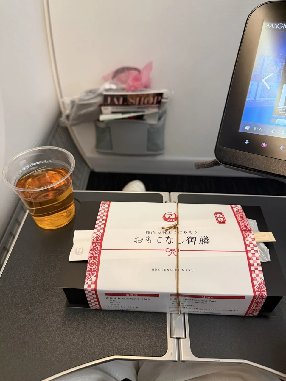 JALの機内食「おもてなし御膳」の箱と、プラカップのお茶。前は壁で、肘掛けから出すモニター