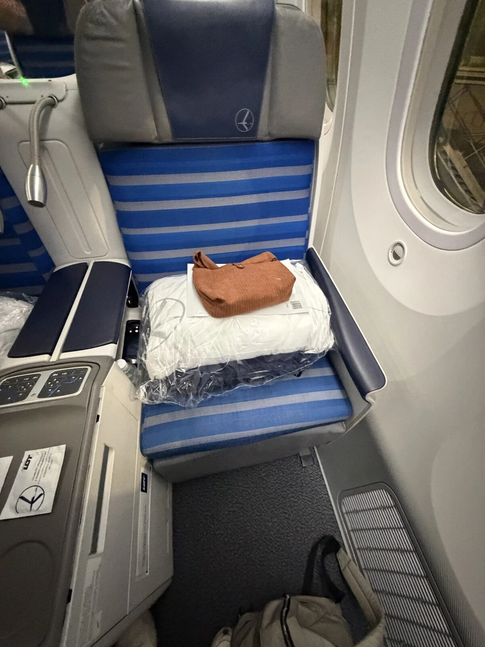 LOT 787ビジネスクラスの座席1A。青と白のストライプのシートに、袋入りの枕と布団、茶色のアメニティポーチ