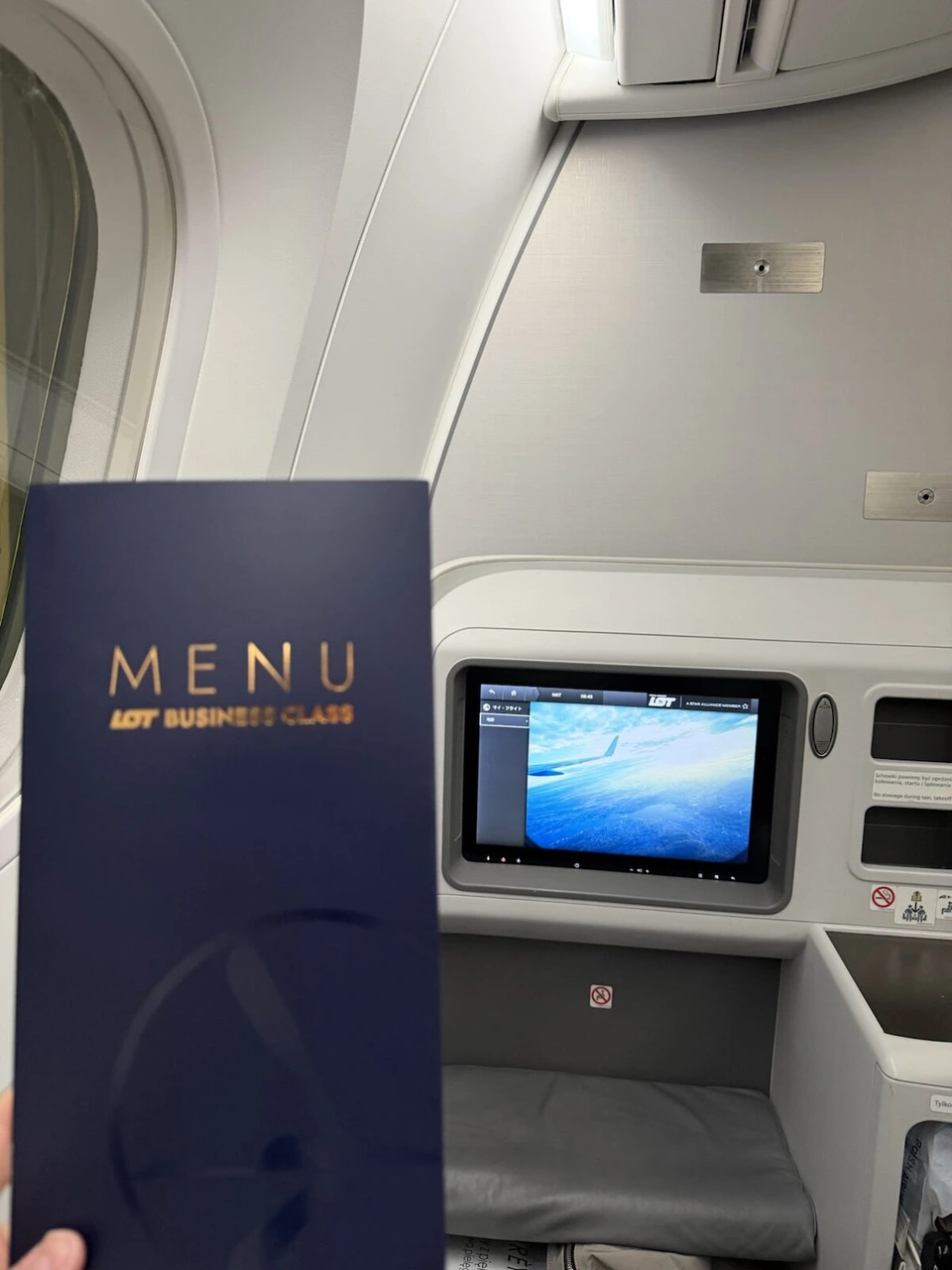 手に持ったLOT BUSINESS CLASSのMENUと、前の壁の機内エンタメの画面