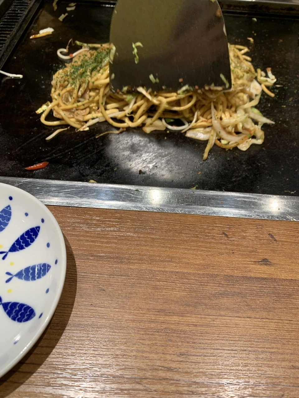 テーブルの鉄板でコテを使って焼きそばを焼いているところ。手前に青い魚の絵の皿