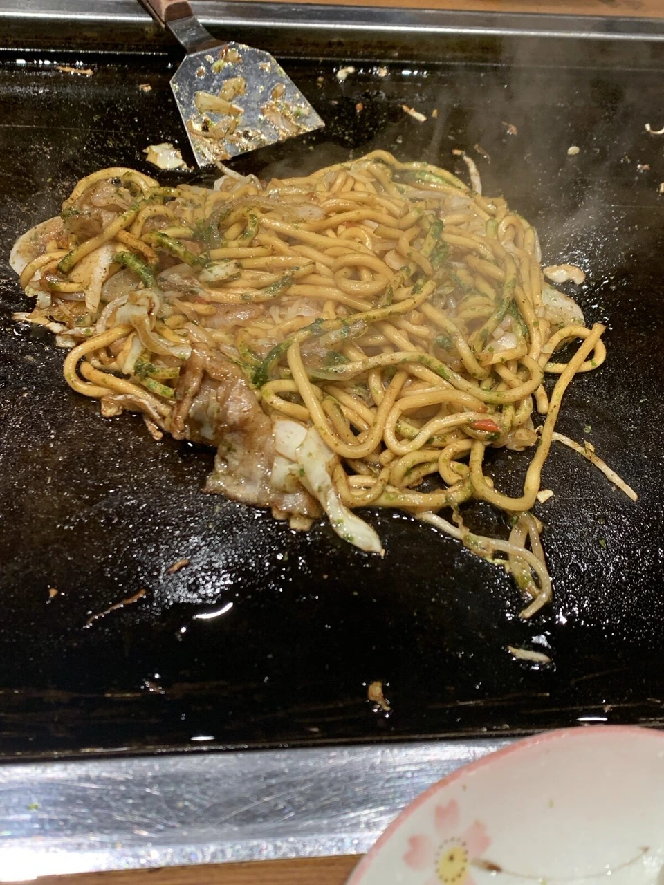 お好み焼道場のテーブルの鉄板で焼いている焼きそば。キャベツと麺に青のりがかかり、奥にコテ