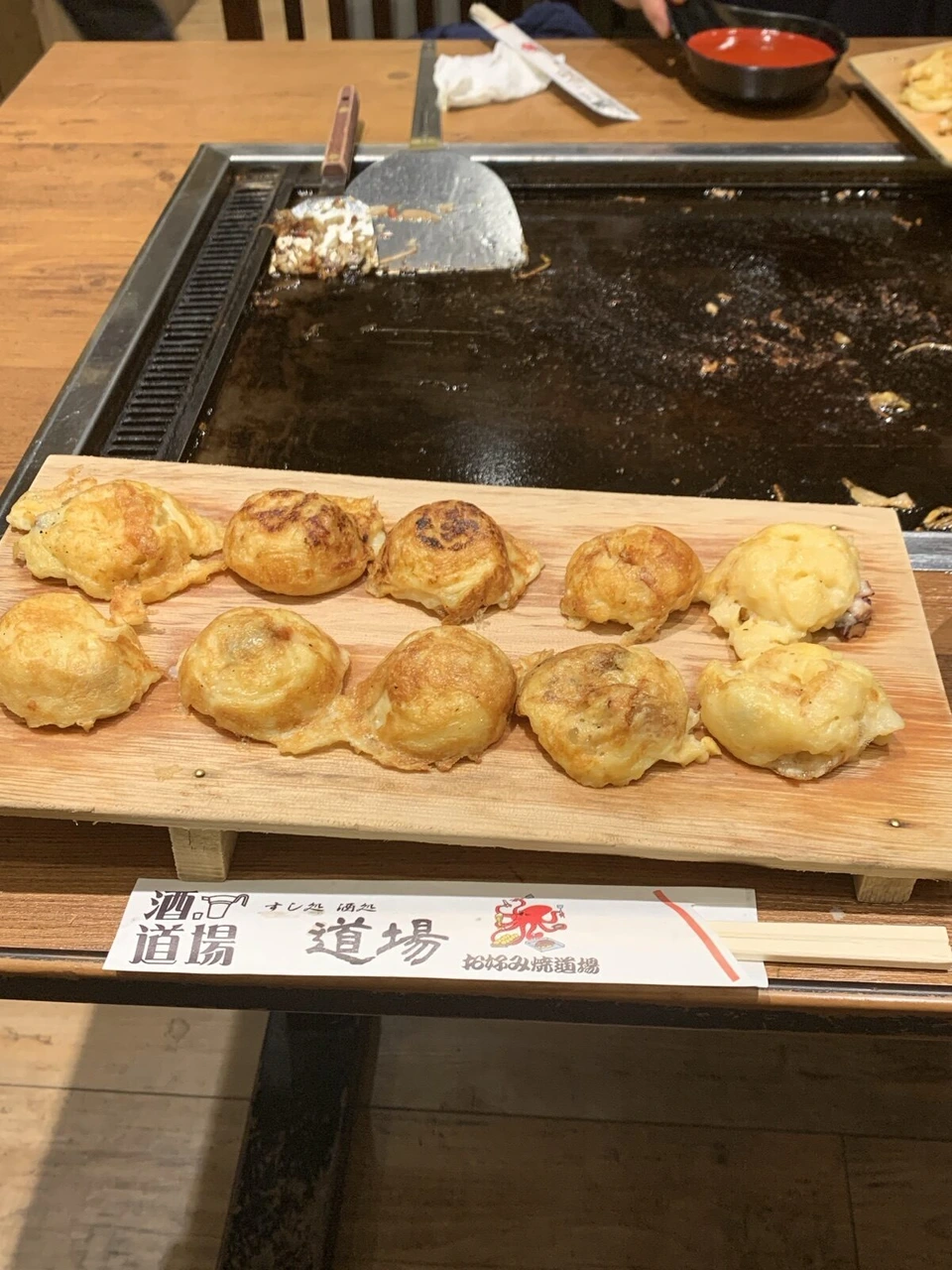 鉄板の手前の木の板に10個並んだ明石焼(玉子焼)と、お好み焼道場・酒道場と書かれた箸袋