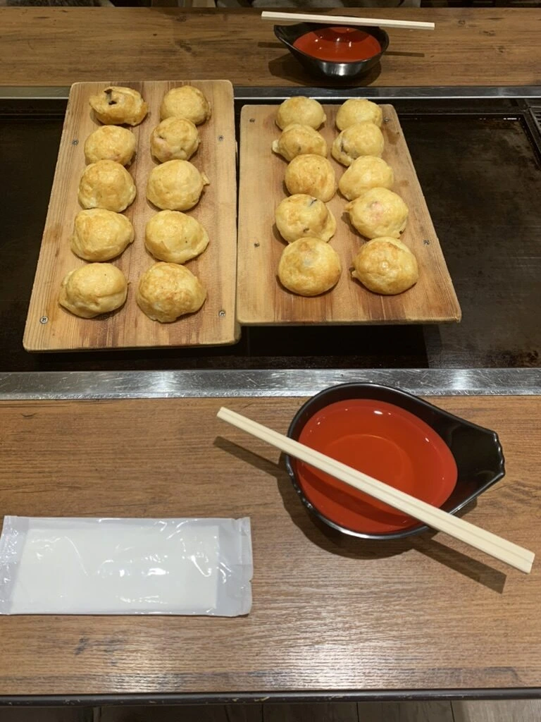 魚の棚商店街の店で出た明石焼。木の板に10個ずつ並んだ玉子焼が2枚と、だしの入った赤い器と箸