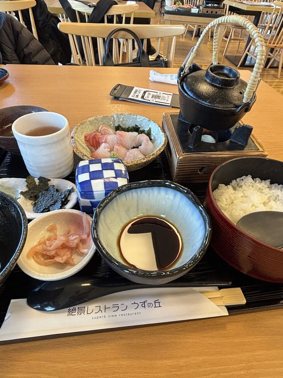 絶景レストラン うずの丘の2025年1月の御膳。鯛とわかめの刺身、たれの器、鉄瓶、ご飯のお椀と箸袋。奥は空席の多い店内