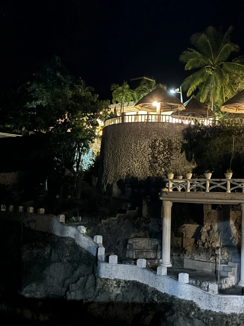 夜の崖の上に建つ白い柱の東屋とライトアップされたヤシの木