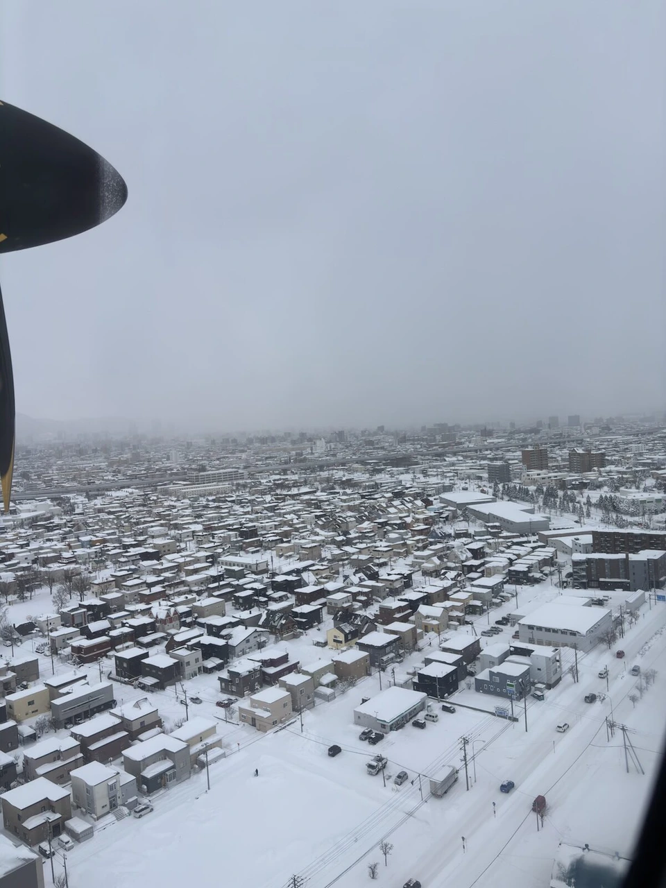 プロペラ機の窓から見た、雪に覆われた札幌の住宅街とプロペラ