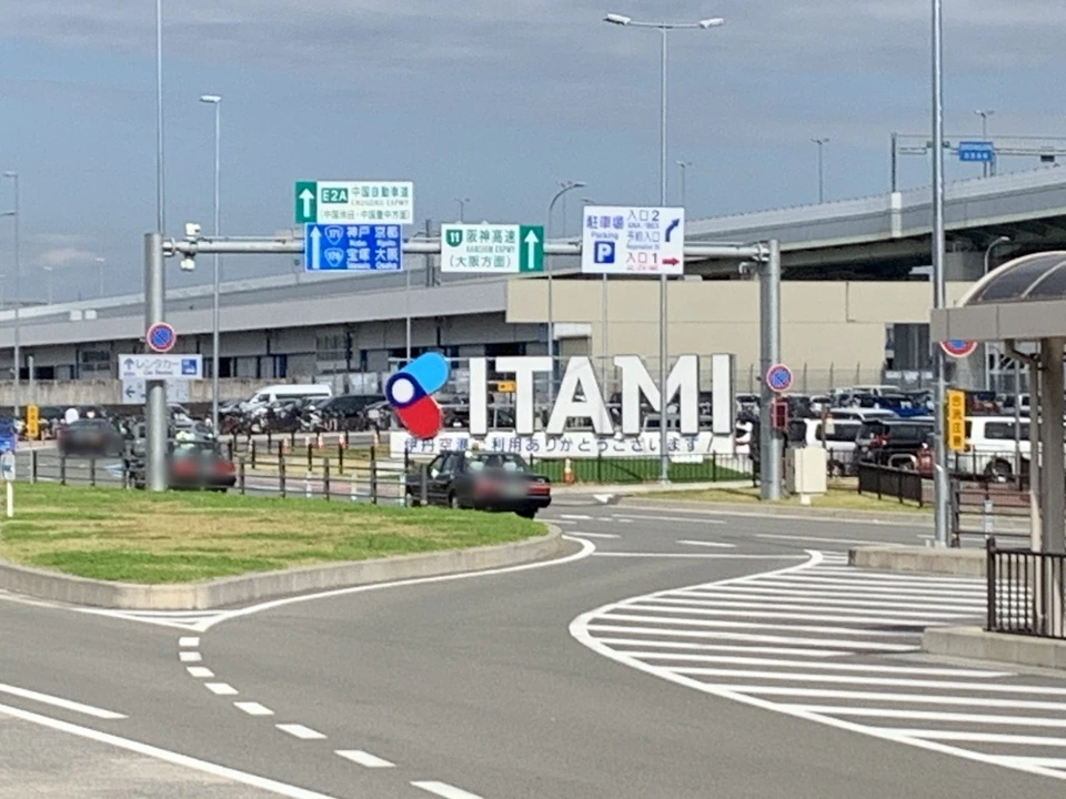 伊丹空港の前の道路と、ITAMIの大きな文字のモニュメント。奥に駐車場と高速道路の案内標識