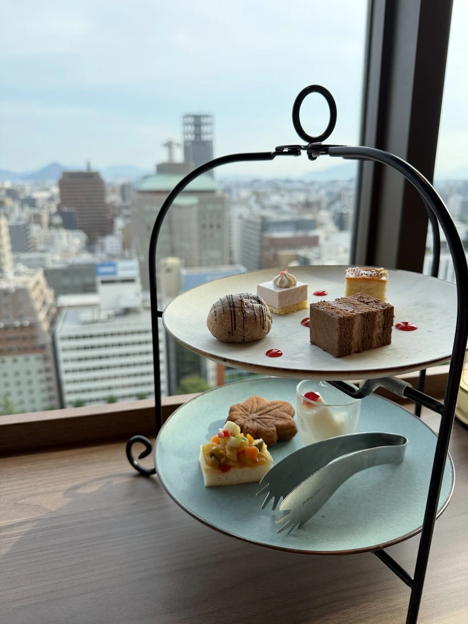 窓際の2段のスタンド。上の段にケーキ、下の段にもみじ饅頭やブルスケッタ、グラスのデザート。窓の外に広島の街