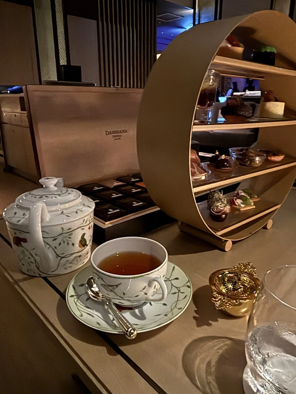 夜のラウンジのテーブル。白いティーポットと紅茶のカップ、奥に丸い金色のスタンドと紅茶の木箱
