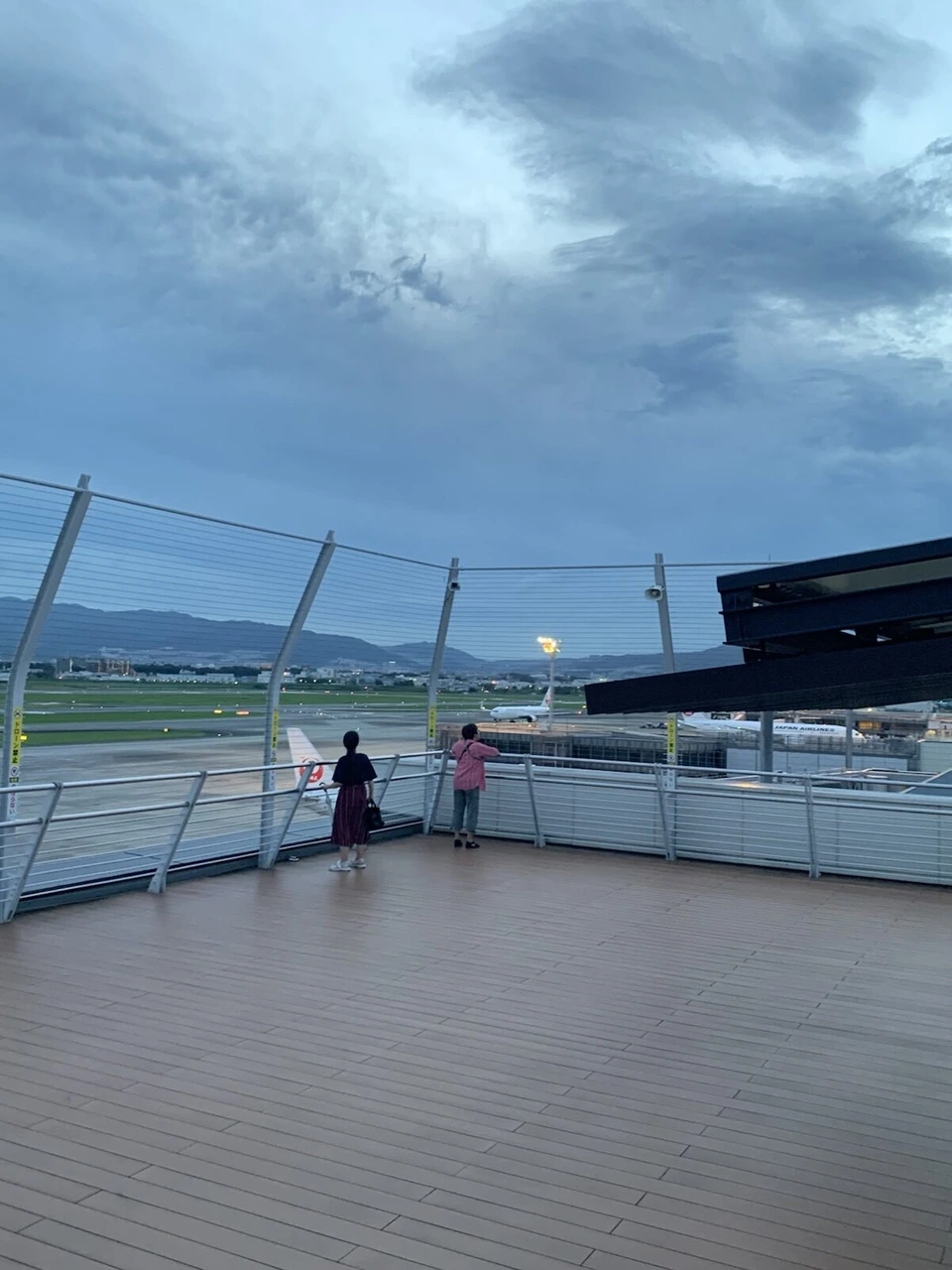 夕暮れの伊丹空港の展望デッキ。ワイヤーの柵の向こうに滑走路と遠くの山並み、空には厚い雲。デッキの奥に人の後ろ姿