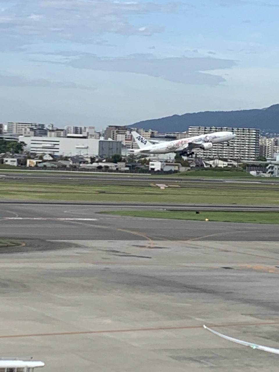 伊丹空港の滑走路から飛び立つ、白いスター・ウォーズ塗装のANA機。奥にマンションと山並み