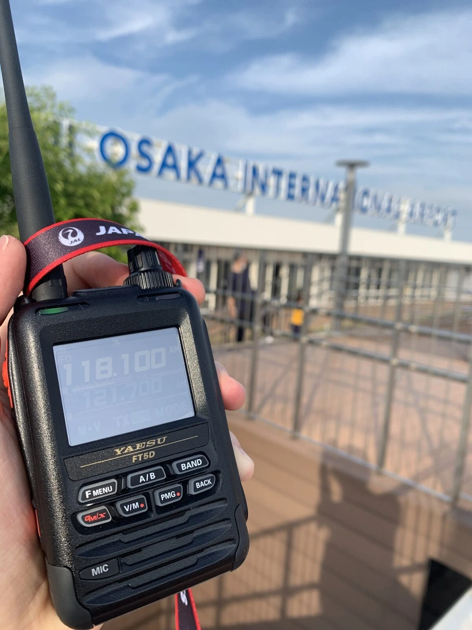 手に持った黒いハンディ型の受信機YAESU FT5D。画面に118.100と121.700の数字、JALのストラップ付き。奥にデッキの柵と空港の文字