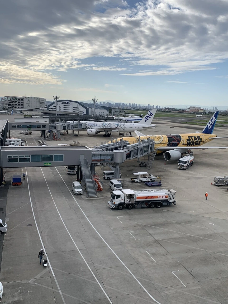 高い位置から見下ろした朝の伊丹空港の駐機場。搭乗橋の向こうに白いスター・ウォーズ塗装のANA機と、黄色いC-3PO塗装のANA機が並び、給油車が止まっている