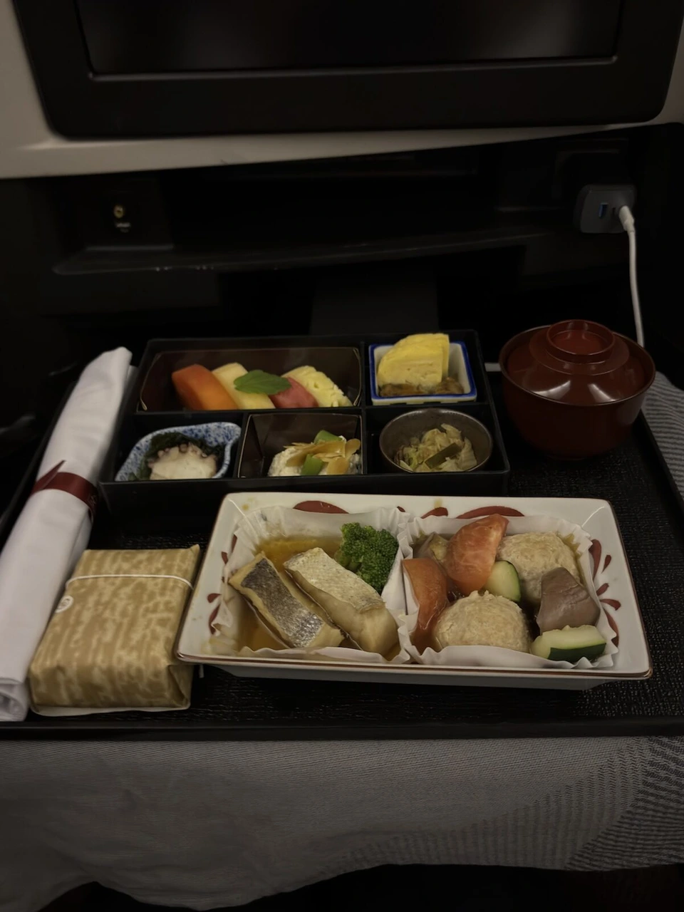 JL724ビジネスクラスの和食の朝食。焼き魚と煮物の皿、小鉢が並んだ箱、汁椀