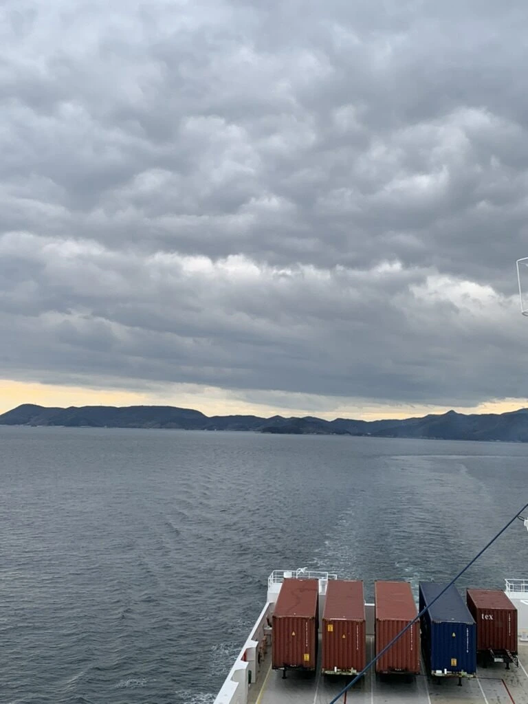 曇り空の下、航跡の先に島影と、後部デッキに積まれたコンテナ