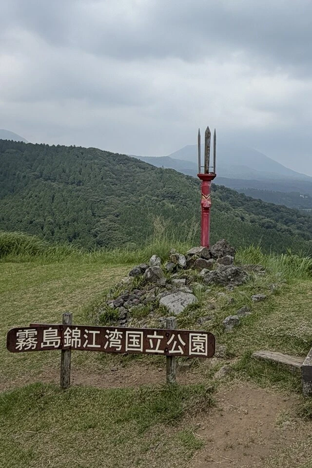 「霧島錦江湾国立公園」の木の看板と、石積みの上に立つ赤い鉾のモニュメント。奥にとがった山