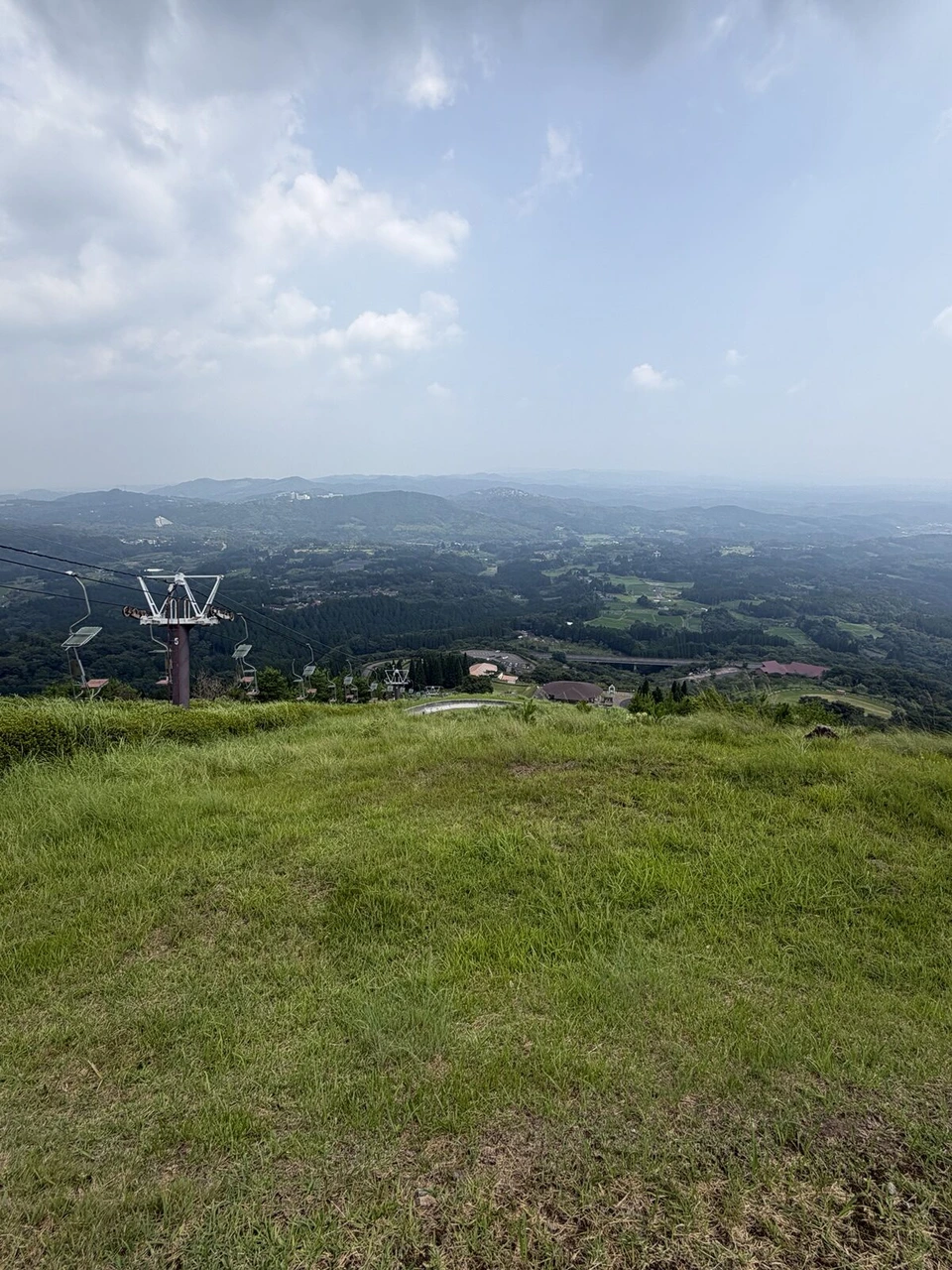 山頂の草地の先に、リフトの赤い鉄塔とふもとの建物、遠くの山並み