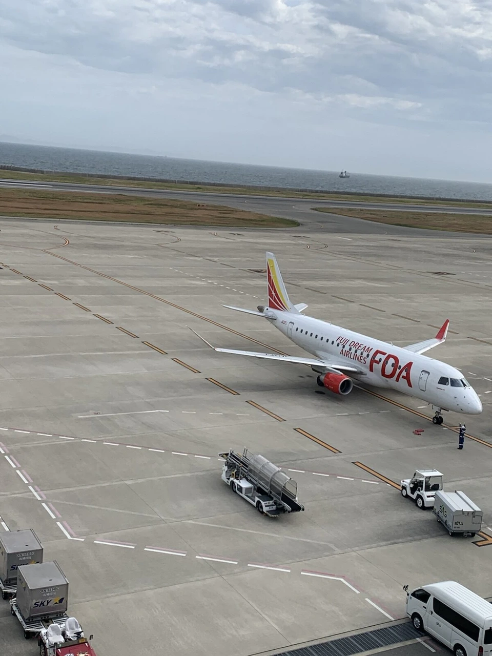 神戸空港の駐機場に止まる、白い胴体に赤い「FDA」の文字と赤い尾翼のエンブラエル機。周りに地上の車両
