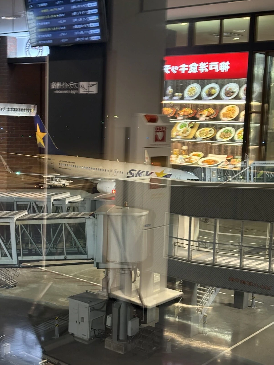 神戸空港のターミナルの中からガラス越しに見たスカイマークの機体。ガラスに、後ろにある飲食店の料理のショーケースと赤い看板が映り込んでいる