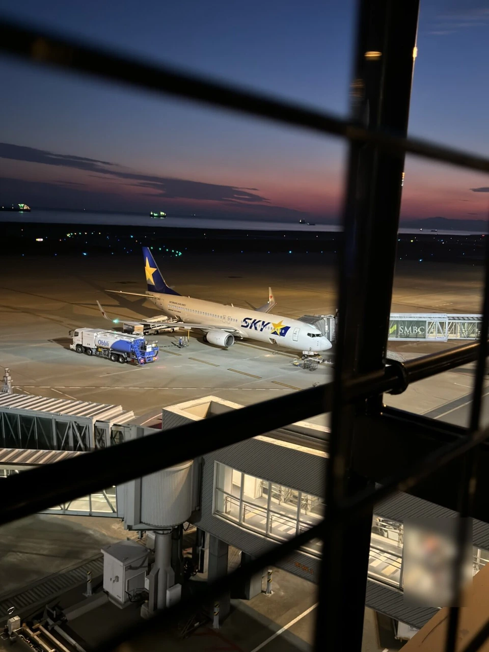 神戸空港の屋上展望デッキの格子の柵越しに見た夕暮れの駐機場。搭乗橋につながるスカイマークの737と、奥に暗くなった海と空