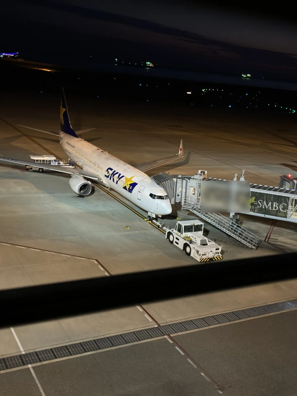 夜の神戸空港で、搭乗橋につながったスカイマークの737。前輪に白いトーイングカーがつながっている。手前にデッキの黒い柵