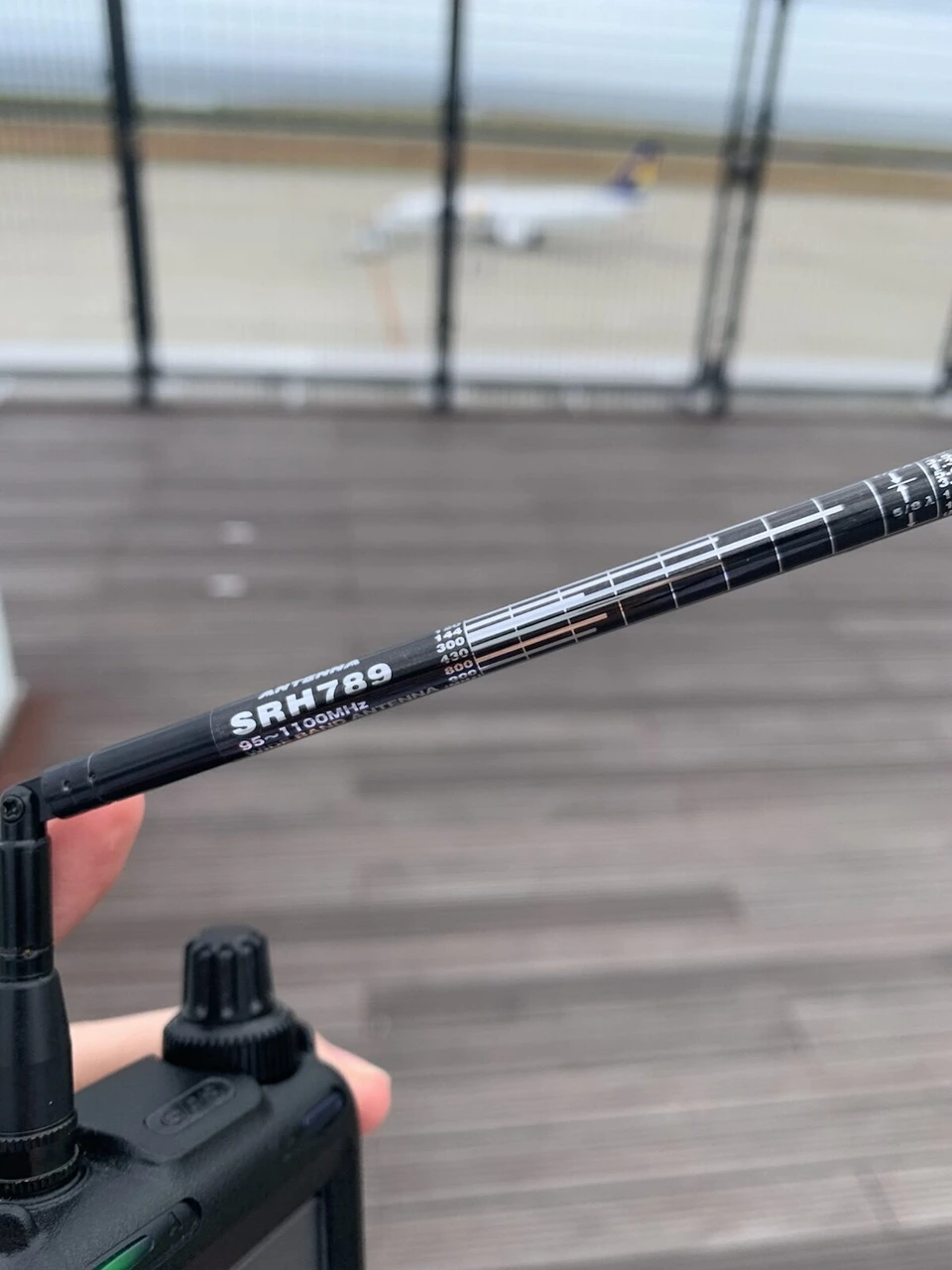 受信機に付けた黒い伸縮式のアンテナ。SRH789の文字が見え、奥にぼやけた飛行機と展望デッキの床