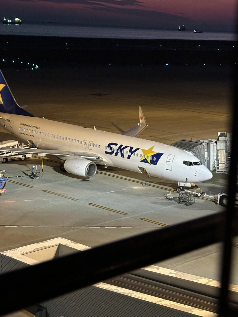 ライトに照らされた、搭乗橋のそばのスカイマークの737。手前に展望デッキの黒い格子が写り込んでいる