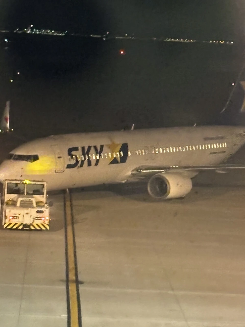 夜の駐機場で、前輪にトーイングカーがつながり搭乗橋から離れたスカイマークの737。ガラス越しに撮ったため、照明の光がぼんやりと映り込んでいる