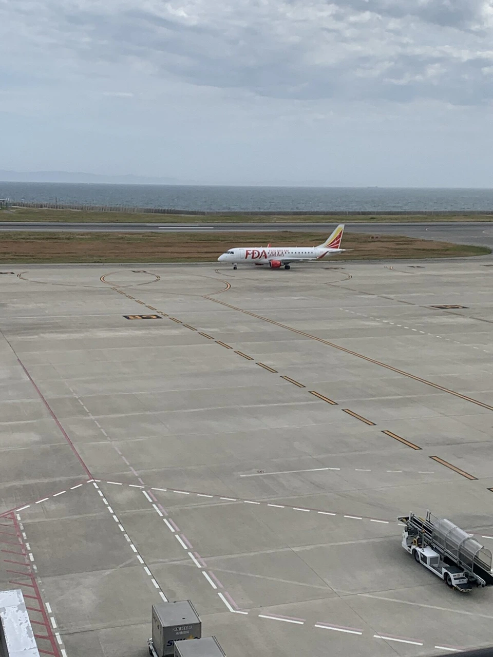 神戸空港の屋上展望デッキから見た広いエプロン。奥の海の手前を、赤い塗装のFDAの小型ジェット機が走っている