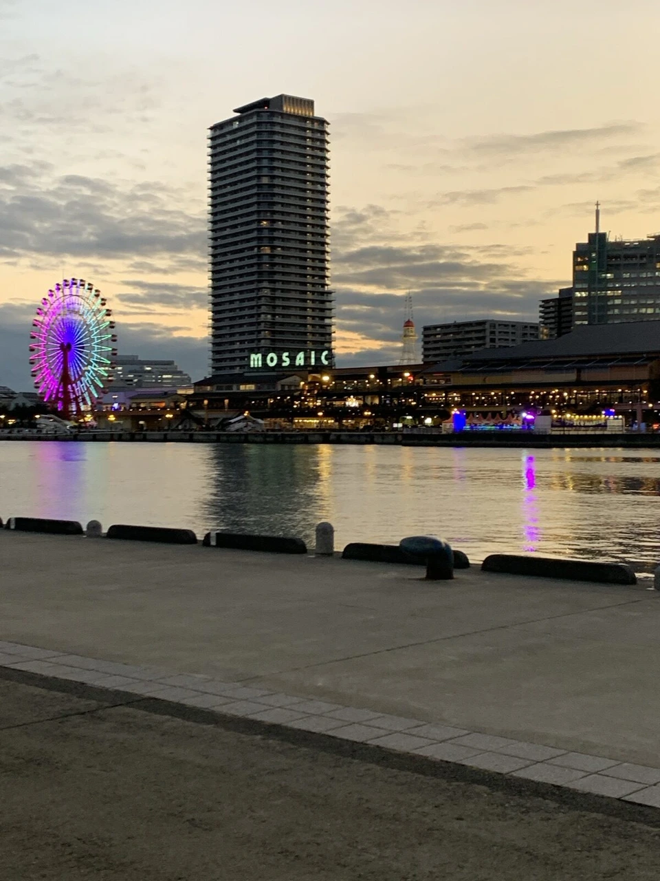 夕焼けの空の下、海の向こうにピンクと紫に光るモザイクの観覧車と高層マンション、MOSAICの文字
