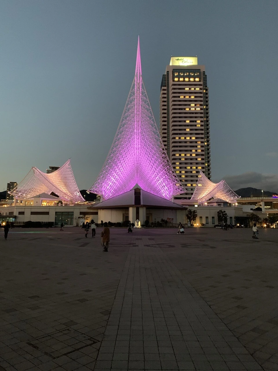 夕暮れの広場の奥で、真ん中の帆がピンクに光る神戸海洋博物館。後ろにホテルオークラ神戸