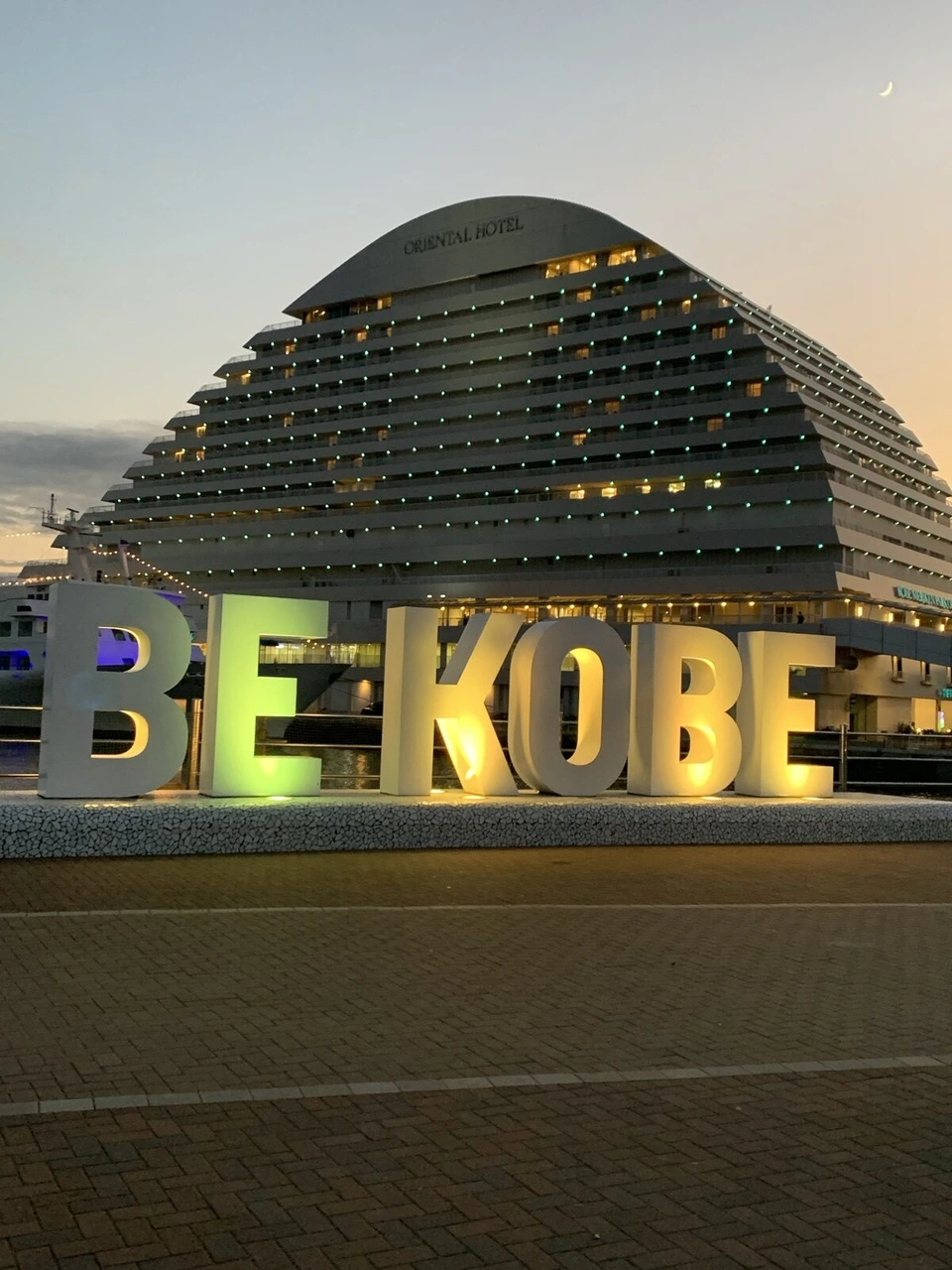 夕暮れのメリケンパーク。黄色や紫に光るBE KOBEの白い文字のモニュメントと、後ろの船の形のオリエンタルホテル。空に細い月