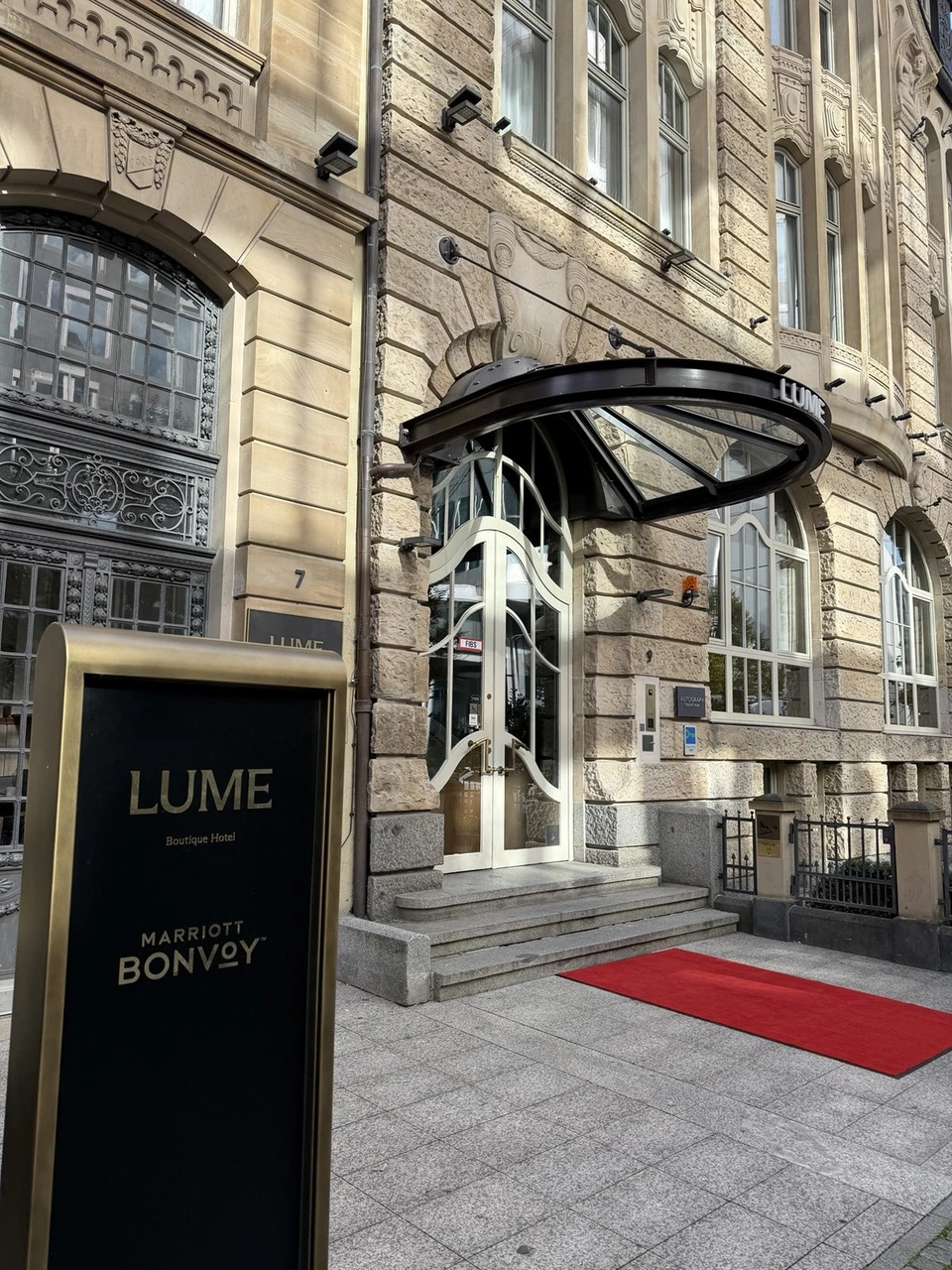 昼のLUME Boutique Hotelの入口。石造りの建物にガラスのひさしと赤いマット、手前にMARRIOTT BONVOYの黒い案内板