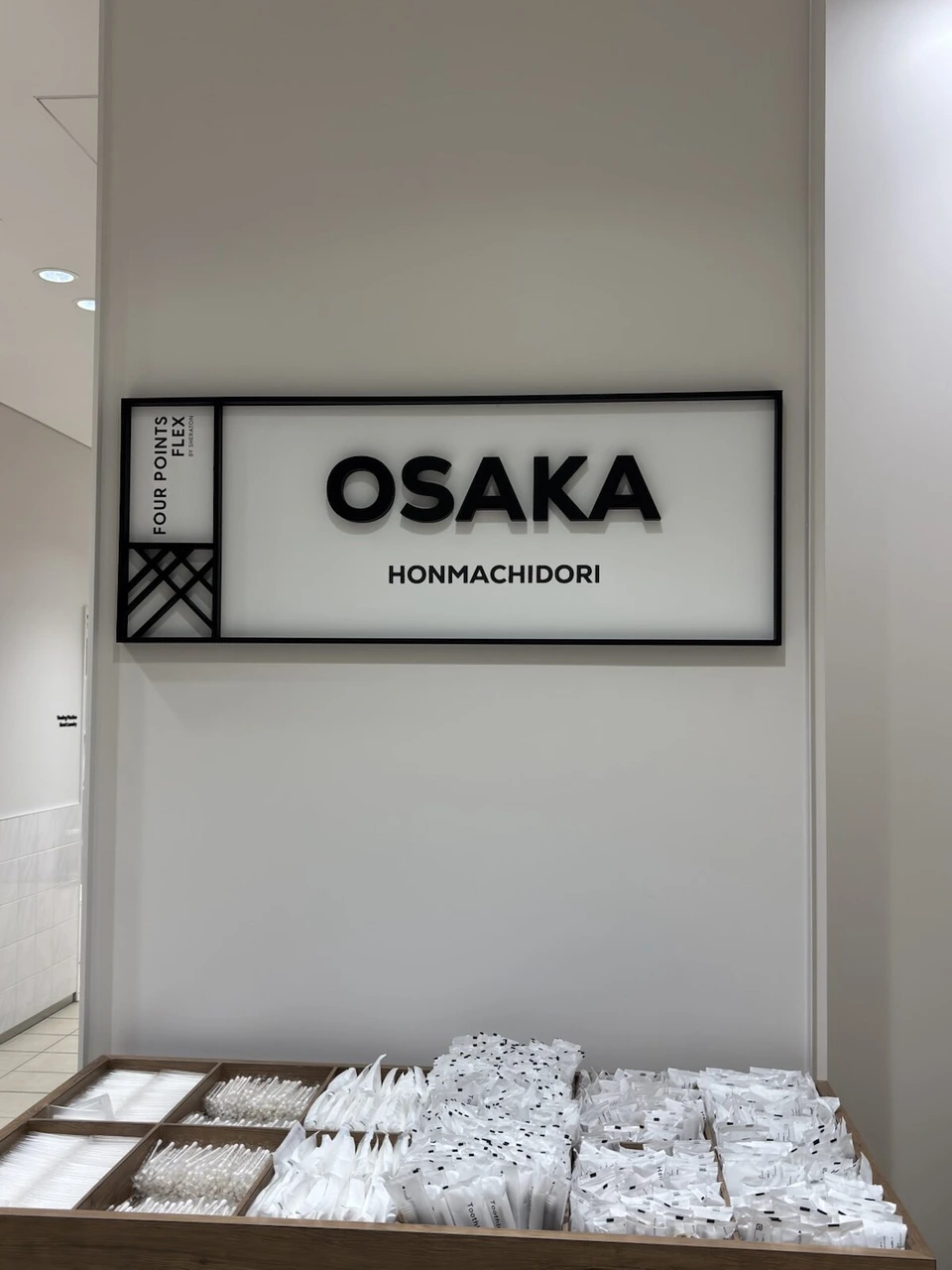フォーポイント フレックス大阪本町のロビー。OSAKA HONMACHIDORIのサインと、白いアメニティが並ぶ棚