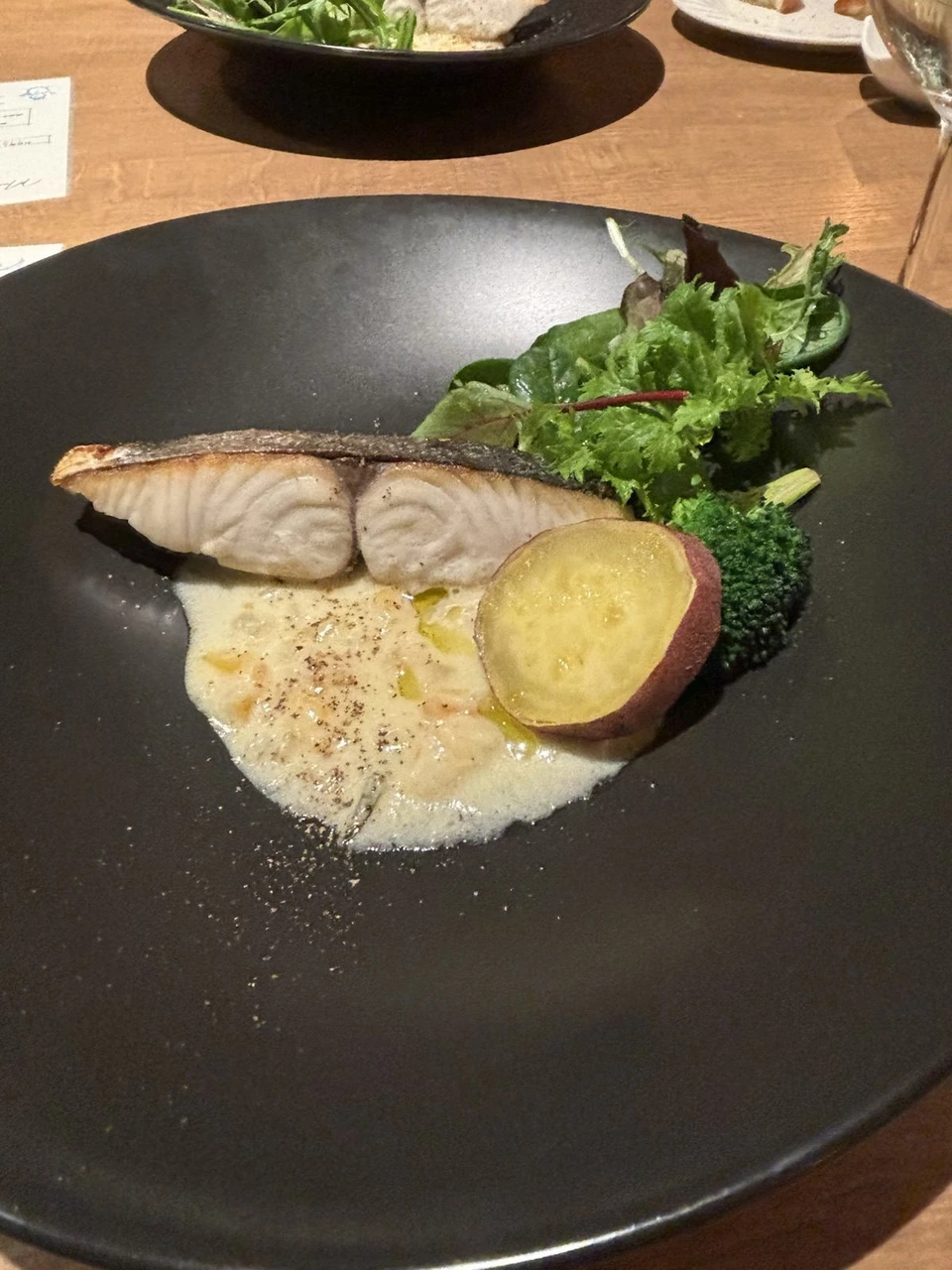 黒い丸皿にのった皮目を焼いた白身魚。クリームのソースに輪切りのさつまいもとブロッコリー、葉野菜が添えてある