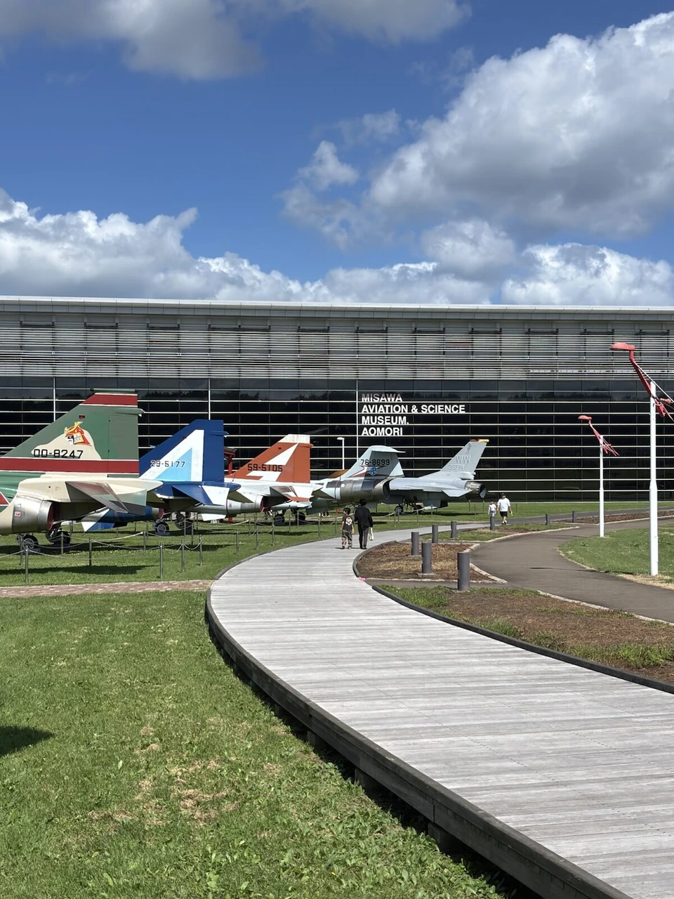 三沢航空科学館の建物の前に、航空自衛隊の戦闘機や練習機が並ぶ屋外展示と木の遊歩道