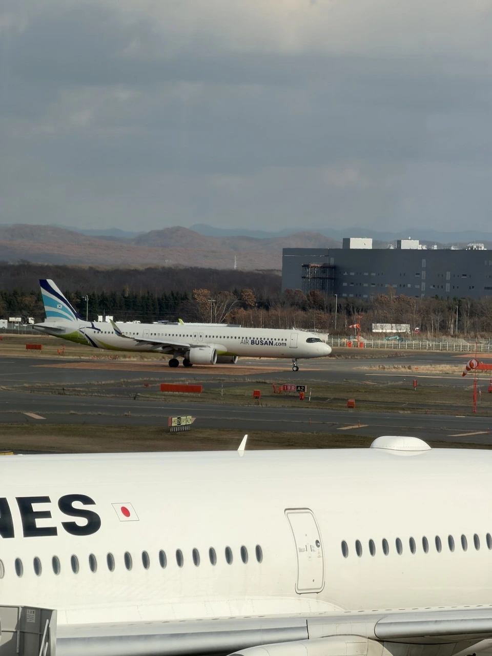 新千歳空港のターミナルの窓から見た誘導路のエアプサンのエアバスA321neo。手前にJAL機の胴体、奥に葉を落とした林と茶色い山