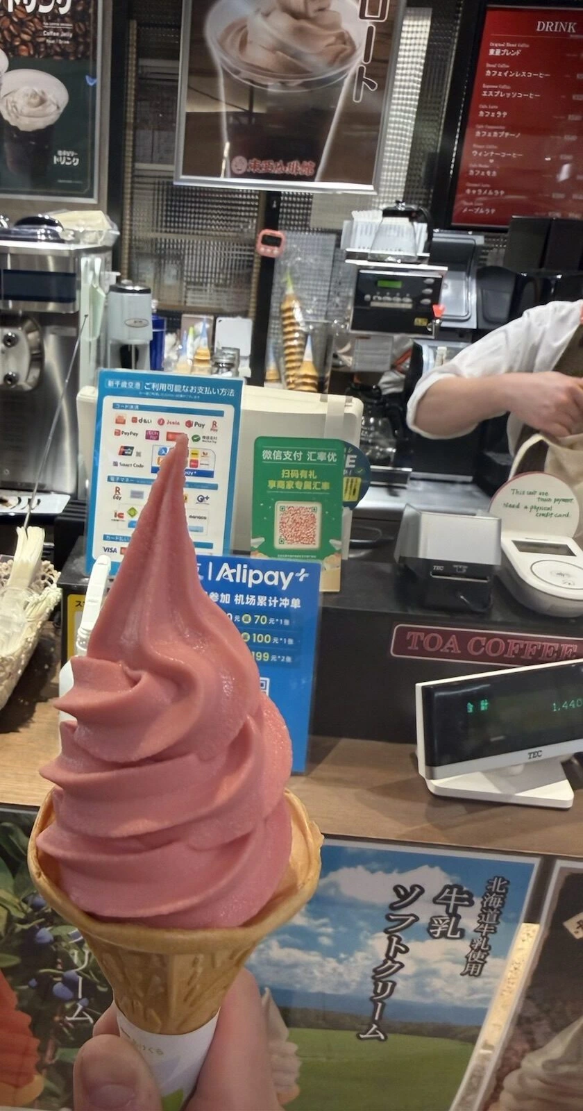 新千歳空港のカフェのカウンターの前で持ったピンク色のソフトクリーム。奥にTOA COFFEEの看板と北海道牛乳ソフトクリームのポスター