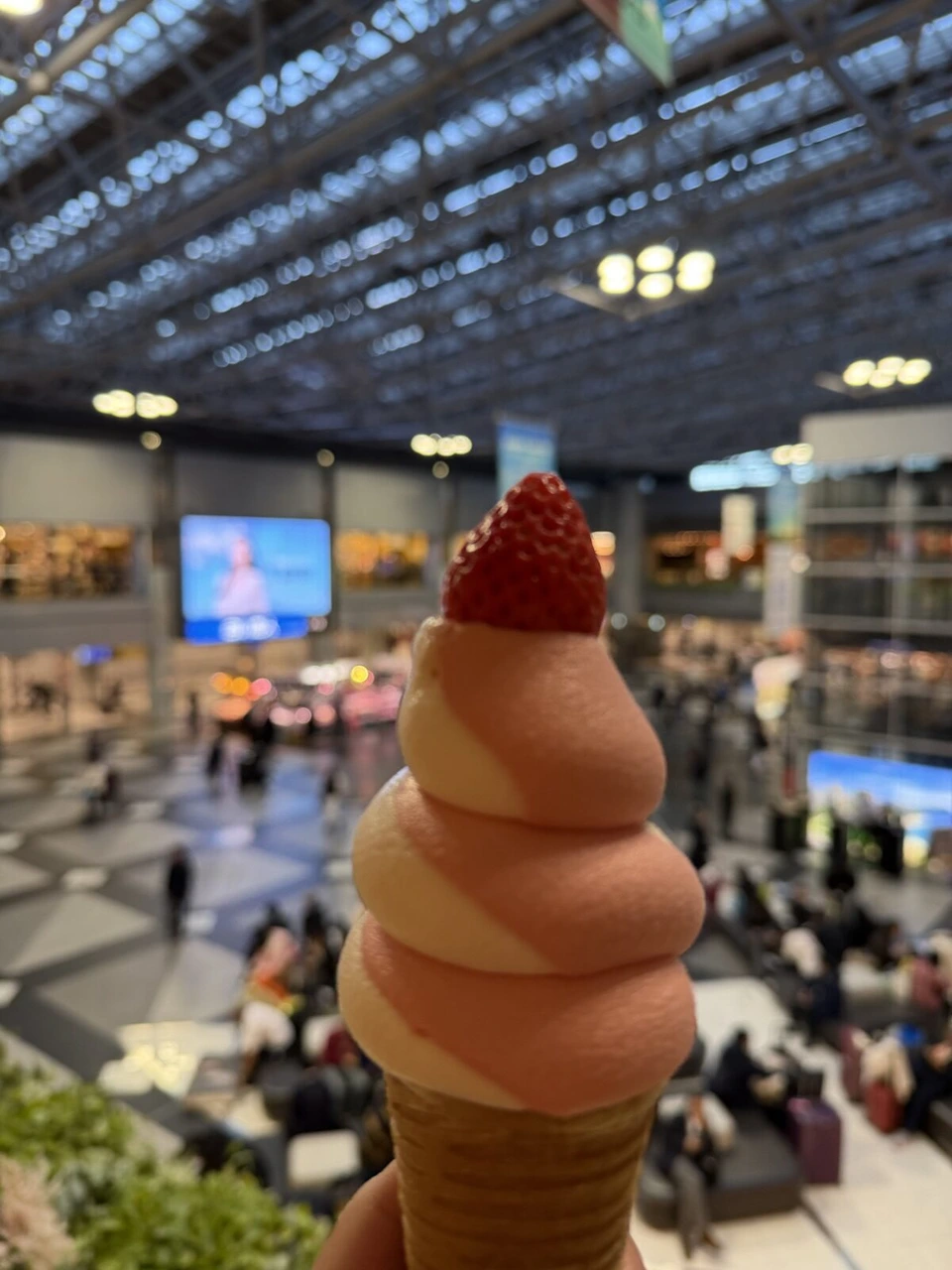 いちごをのせたピンクと白のうず巻きのソフトクリーム。背景にぼやけた新千歳空港の吹き抜けのセンタープラザ