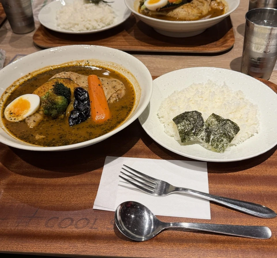 木のトレーにのったスープカレーとごはん。スープの中に鶏肉、ゆで卵、にんじん、ブロッコリー、かぼちゃ、なす。ごはんにはのりが2枚。奥にもう1皿のスープカレー