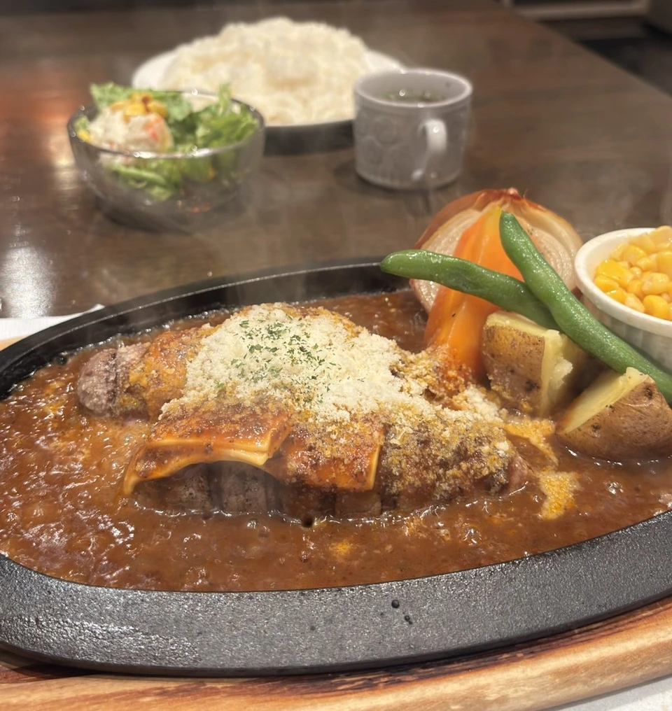 鉄板にのったハンバーグ。チーズと粉チーズがかかり、デミグラスソースから湯気が立つ。付け合わせはじゃがいも、にんじん、玉ねぎ、いんげん、コーン。奥にサラダとスープ、ごはん